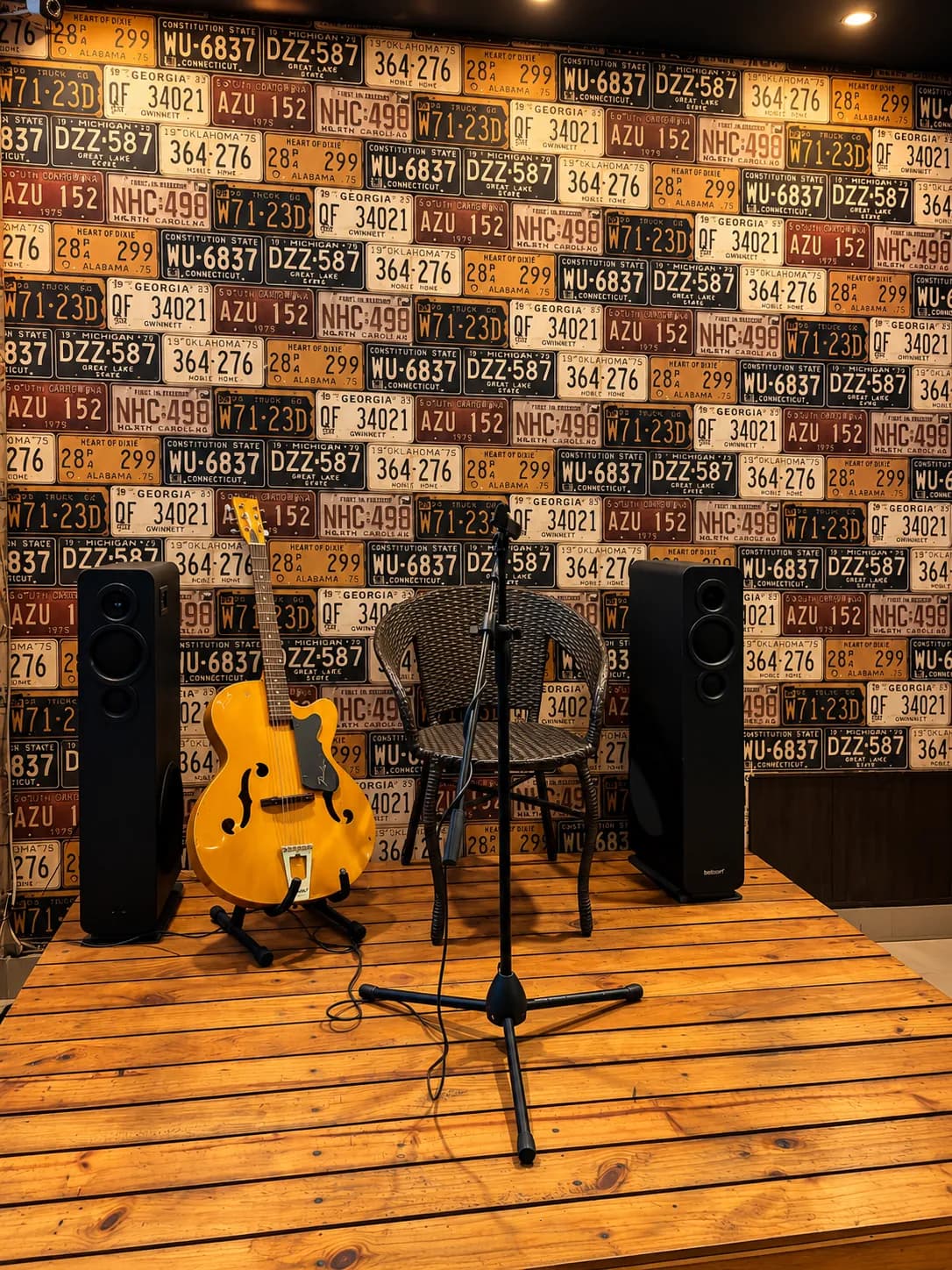 A small wooden stage with a guitar, mic and two speakers in front of a wall of vintage licence plates